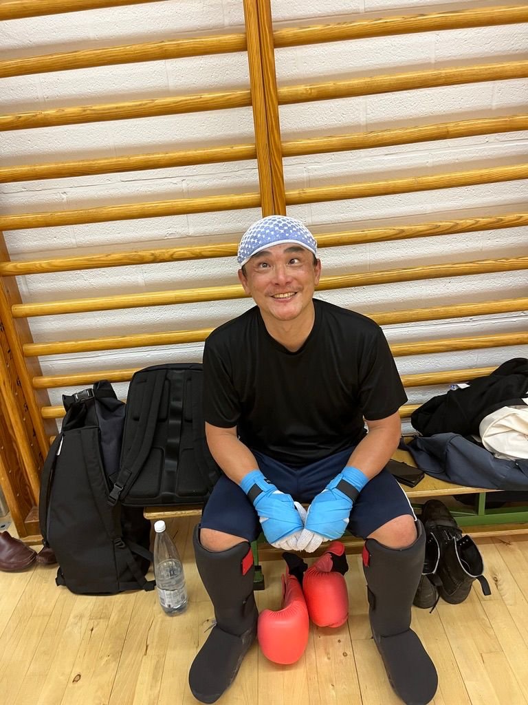 Homme assis sur un banc en bois avec des équipements de sport, portant un bonnet, des gants bleus, des protège-tibias et des gants, dans une salle de sport ou un espace d'entraînement.