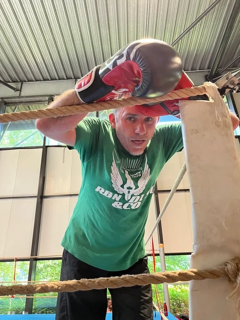 Un homme portant des gants de boxe noirs et rouges, en position de garde, dans une salle de boxe ou un gymnase, regardant vers le bas avec une expression concentrée.