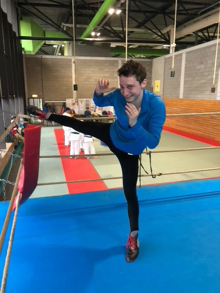 Une personne en tenue de sport effectue une figure de boxe française dans un gymnase avec un ring, des autres pratiquants et du matériel sportif en arrière-plan.