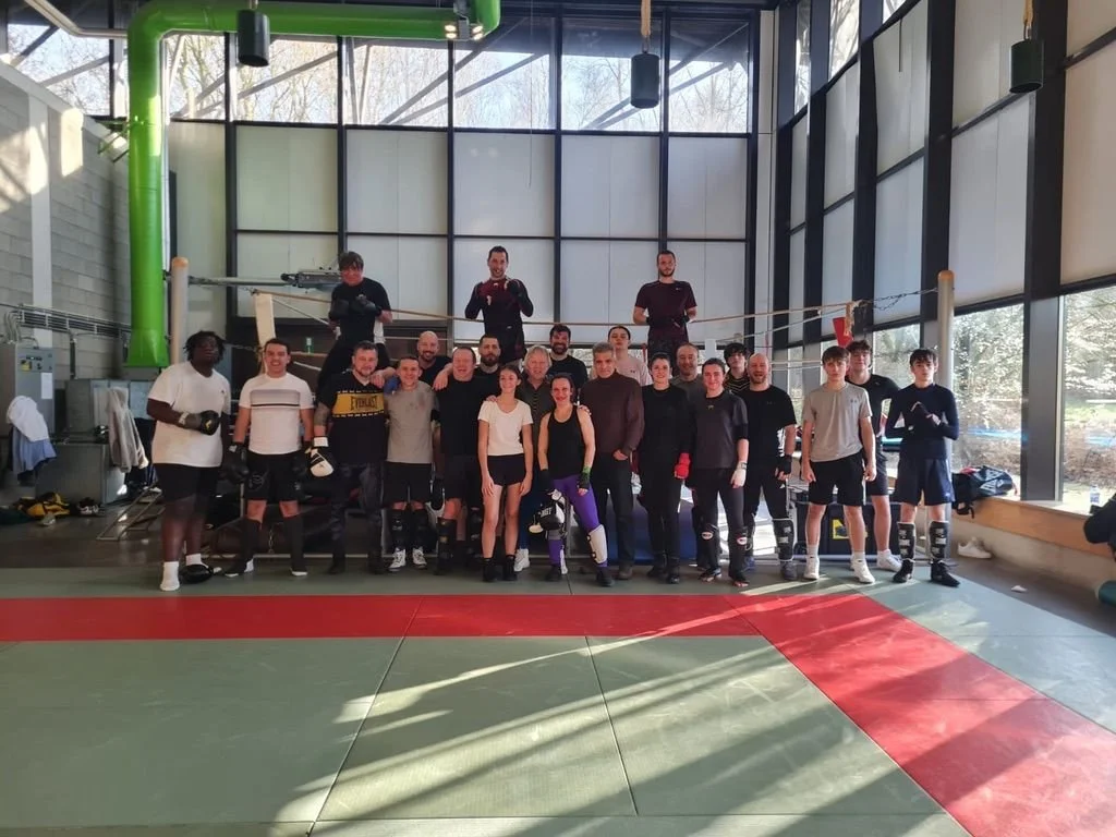 Groupe de personnes dans un centre d'entraînement de boxe, certains portant des gants de boxe et des protections, devant un ring de boxe.