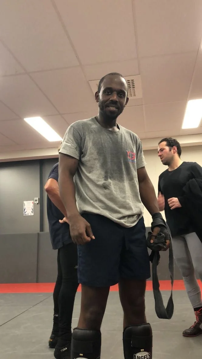 Homme souriant en tenue de sport dans une salle d'entraînement, portant des protections aux jambes et tenant des gants ou équipement de boxe, avec deux autres personnes en arrière-plan.