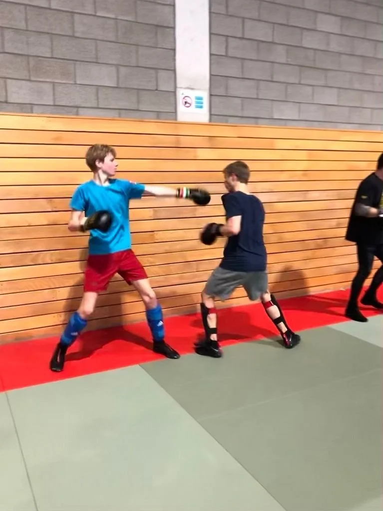 Deux jeunes garçons pratiquant la boxe en compétition, portant des gants de boxe, l'un en t-shirt bleu et short rouge, l'autre en t-shirt noir et short gris