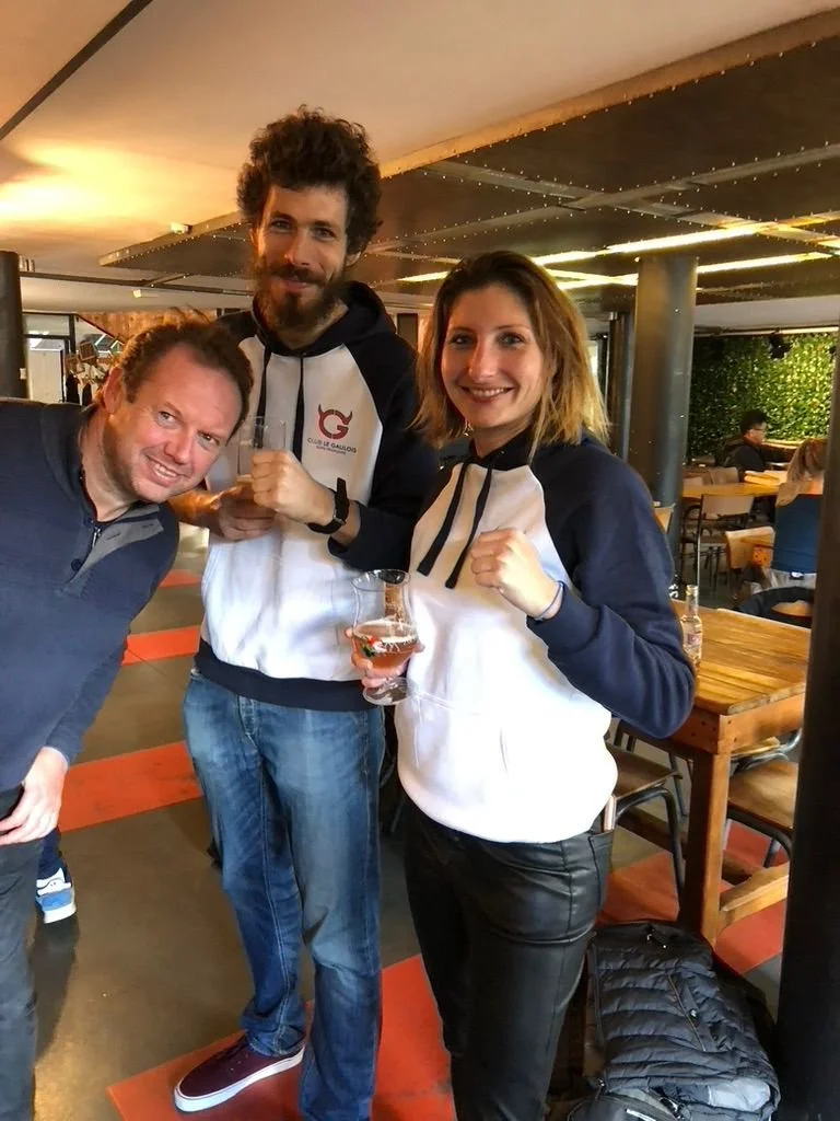 Trois personnes posant pour une photo dans un restaurant ou bar, deux hommes et une femme, l'homme au milieu tient un verre et la femme a un verre à la main. Ils sourient et portent des vêtements décontractés, avec un décor intérieur chaleureux.