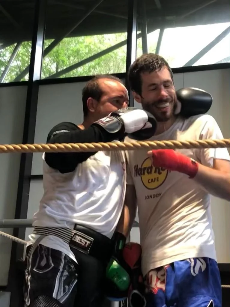 Deux hommes de boxe en tenue sportive, souriants et amis, posent après un entraînement dans un gymnase, avec parapluie, dans un environnement moderne avec grandes fenêtres.