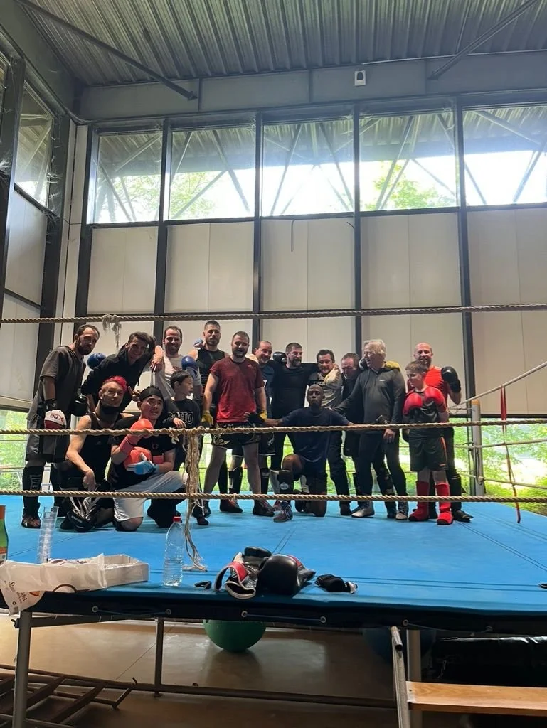 Groupe de personnes de boxe dans un gymnase avec ring de boxe, quelques sacs de boxe, et équipement de sport.