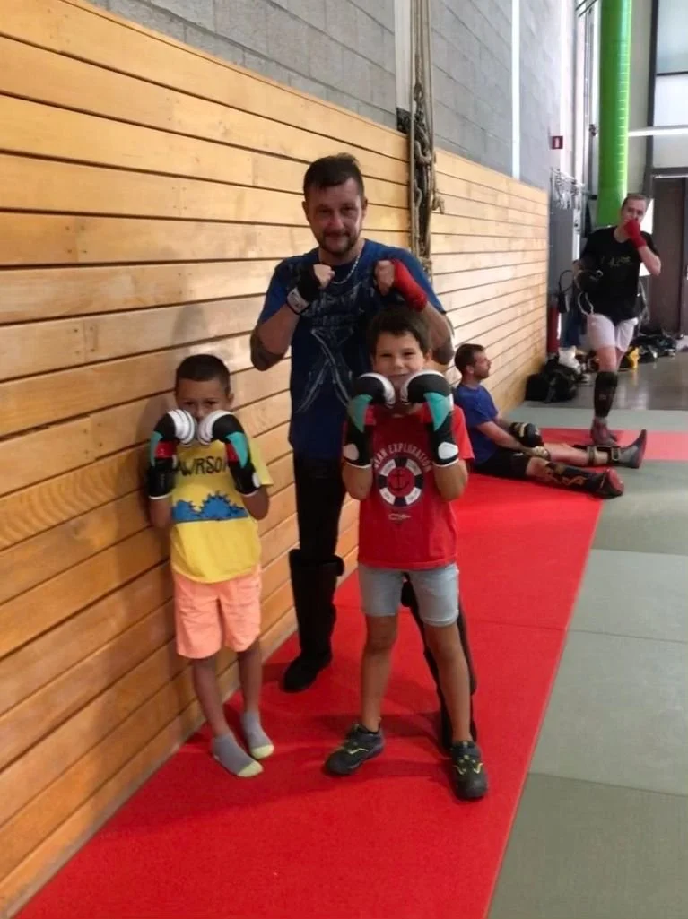 Deux jeunes garçons en équipement de boxe, posant avec un entraîneur, à côté d'une femme en arrière-plan dans une salle d'entraînement. Les garçons portent des gants de boxe et sourient, l'un en jaune et l'autre en rouge.