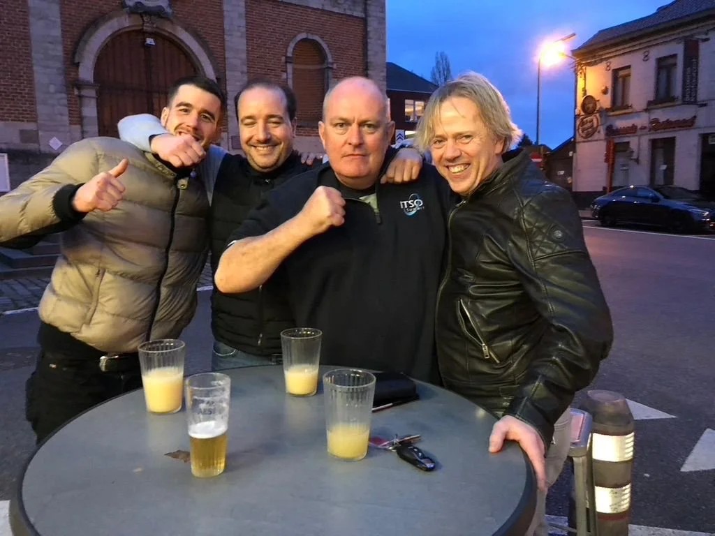 Quatre hommes souriants posent ensemble à une table en plein air, avec trois verres de jus et une bière."
