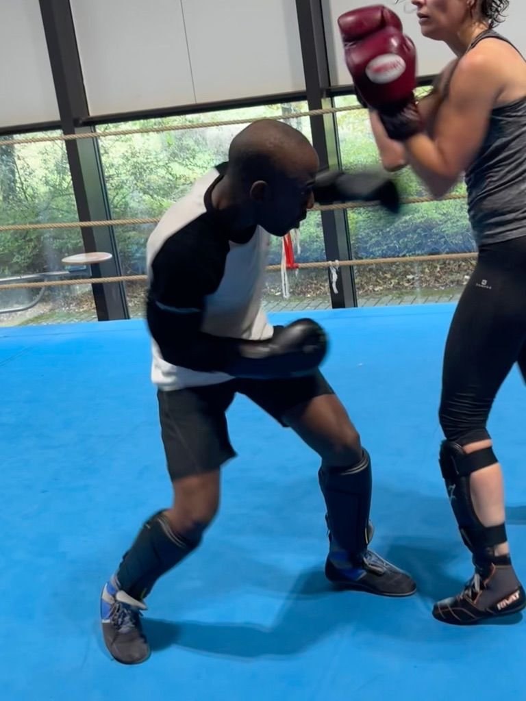Deux boxeurs s'entraînent dans un gymnase, l'un frappe l'autre qui porte des protections et des gants de boxe.