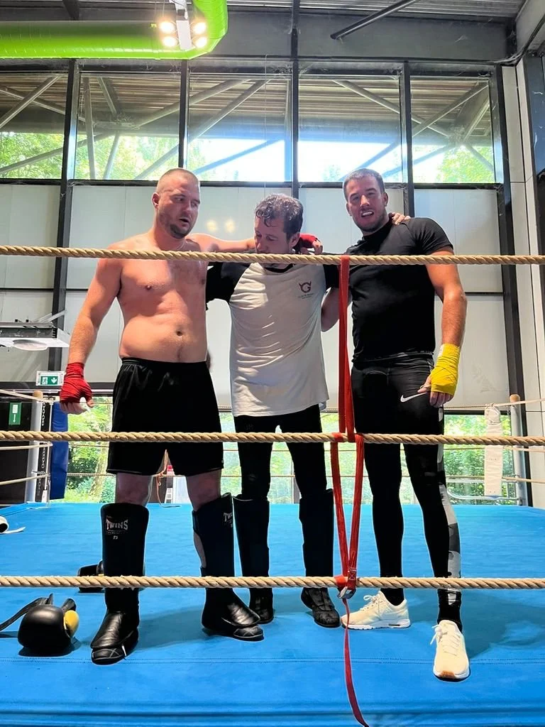 Trois boxeurs dans un ring, deux en t-shirt et l'autre en short, portant des gants de boxe, l'un semble blessé ou fatigué, avec un coach ou un assistant entre eux.