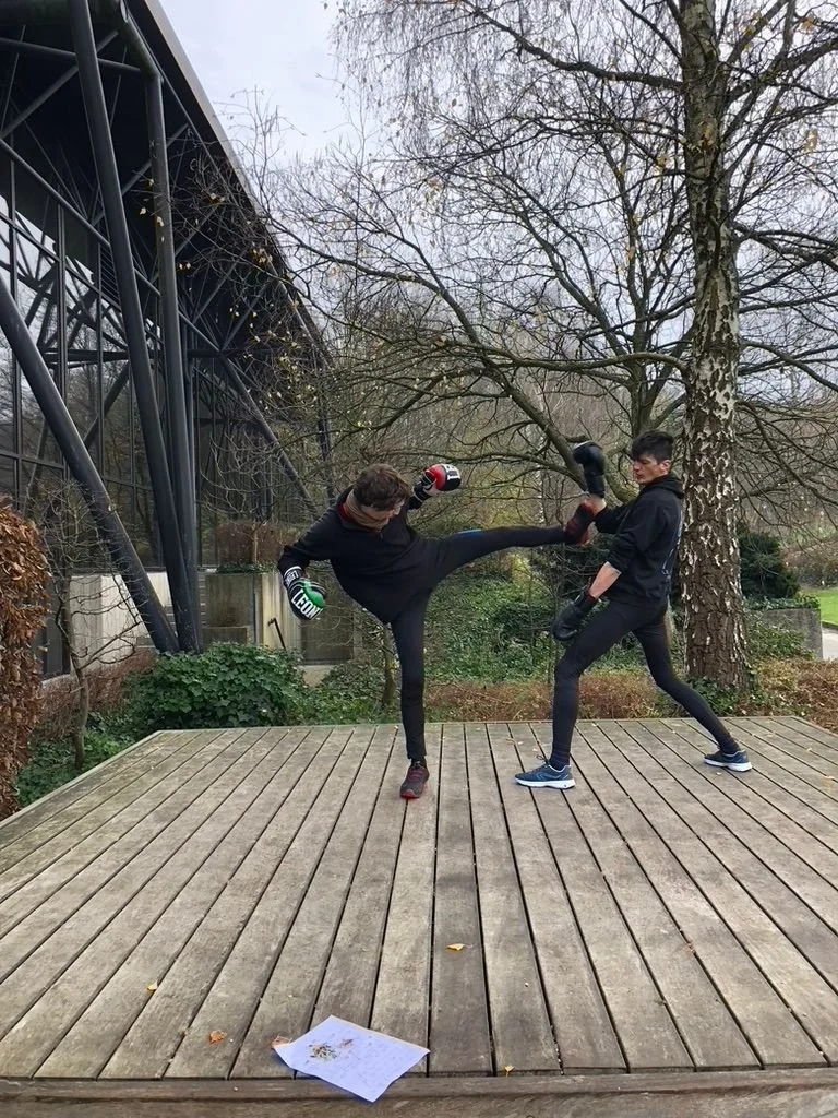 Deux personnes en tenue de sport pratiquent la boxe française, l'une donne un coup de pied à l'autre sur une terrasse en bois, avec des arbres et un bâtiment en arrière-plan.