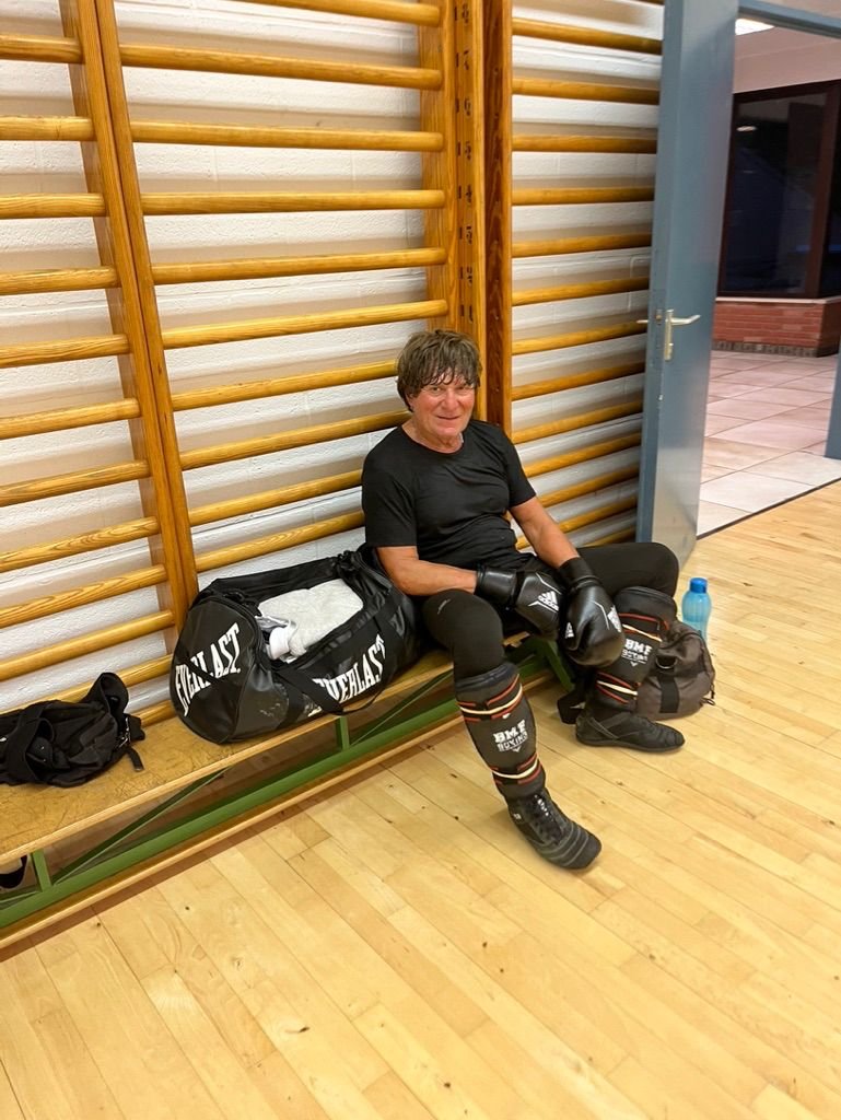 Un homme en tenue de boxe assis sur un banc dans une salle de sport, avec un sac de sport et une bouteille d'eau à côté.