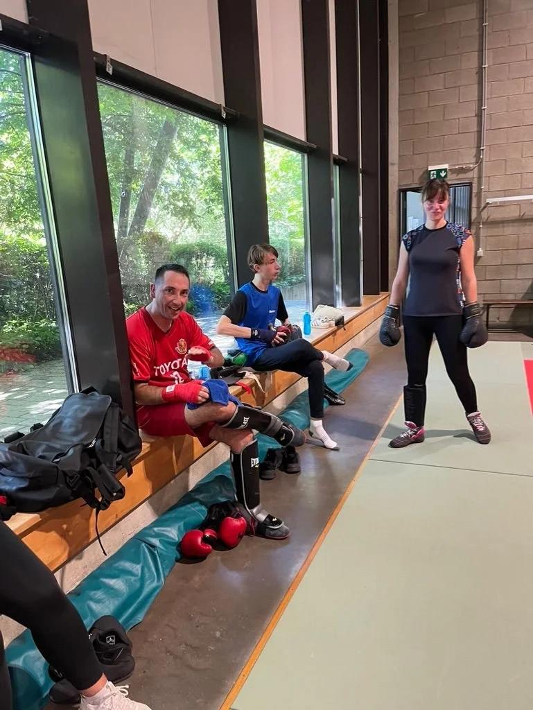 Trois personnes en vêtements de boxe, deux assises sur un banc et une debout dans une salle de sport, avec des gants de boxe et des sacs de boxe à proximité.