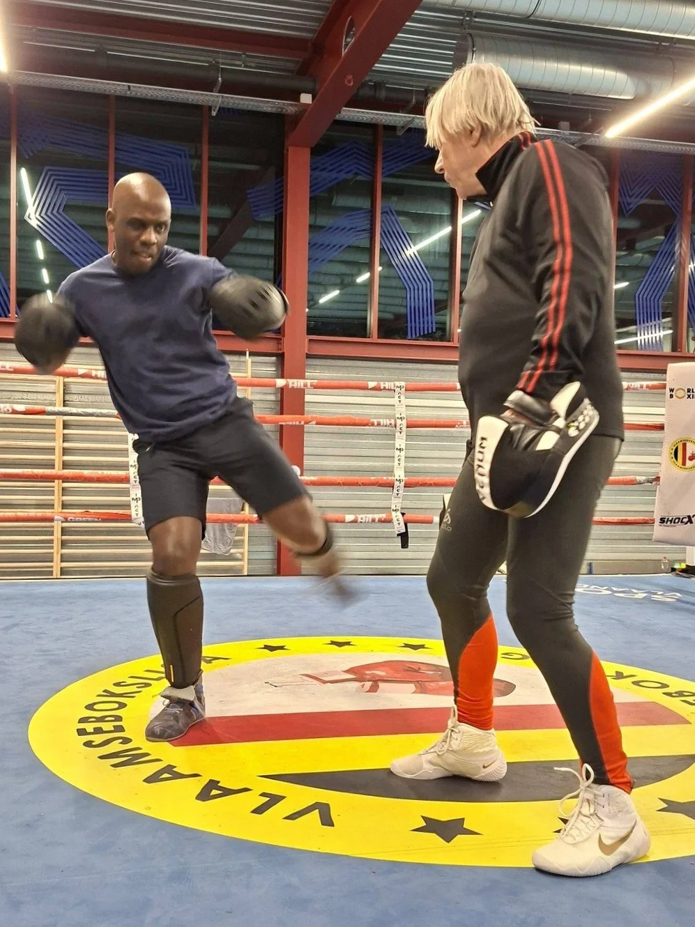 Deux hommes s'entraînent à la boxe dans un gymnase avec un ring, l'un est en train de frapper, l'autre observe, tous deux portent des équipements de boxe.