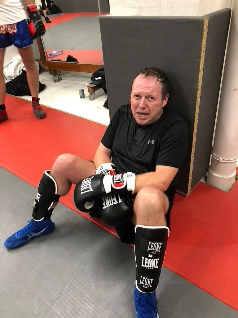 Un homme en tenue de boxe, avec des protège-tibias et des gants, assis par terre dans une salle d'entraînement, ayant l'air épuisé.