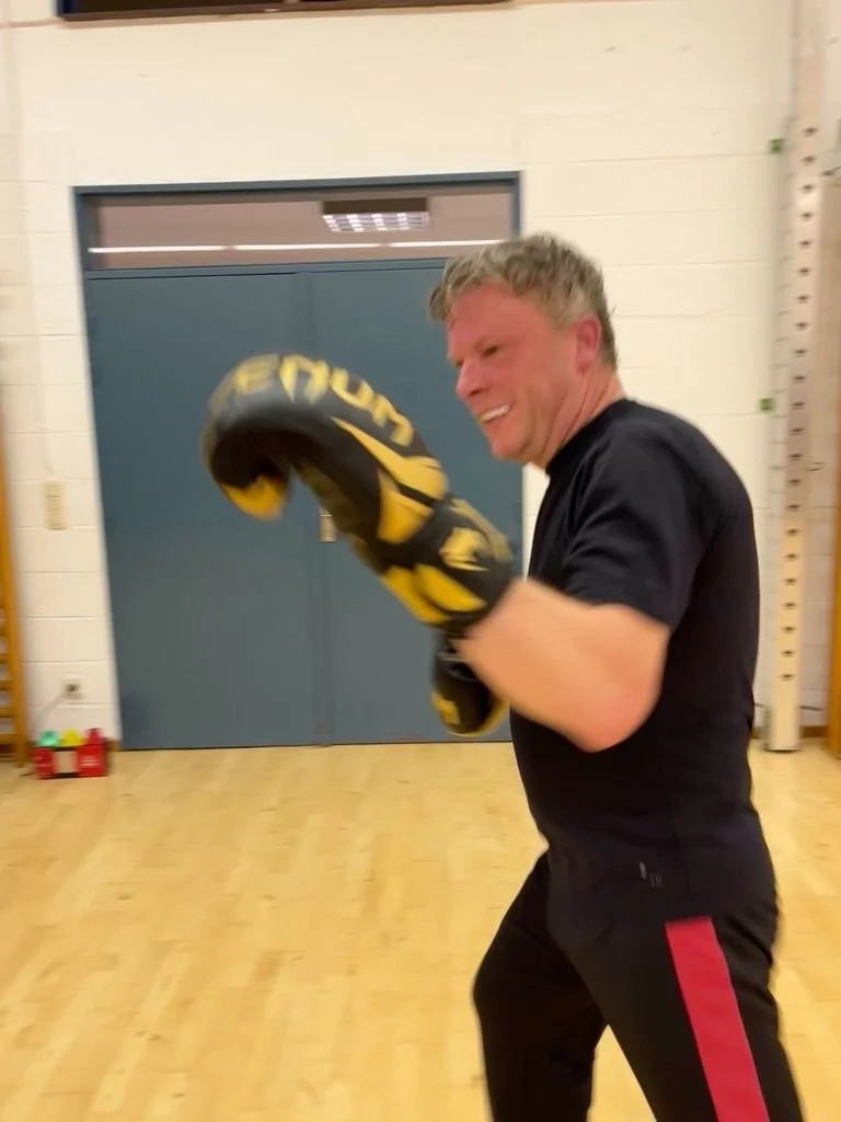 Un homme en train de faire de la boxe, portant des gants de boxe noirs et jaunes, dans une salle d'entraînement, souriant.