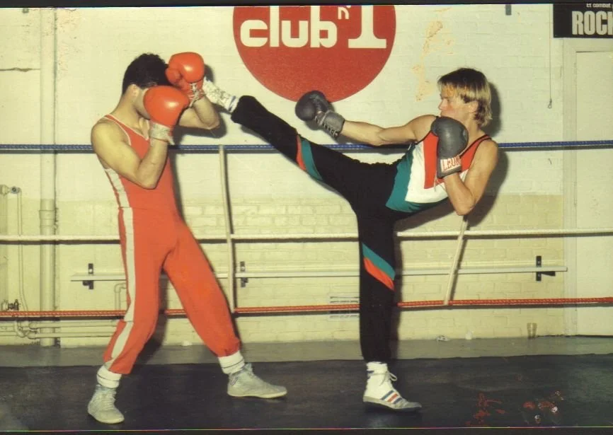 Deux jeunes boxeurs s'entraînent dans un gymnase de boxe. L'un pratique un coup de pied de haute volée sur l'autre, qui se protège avec des gants de boxe.