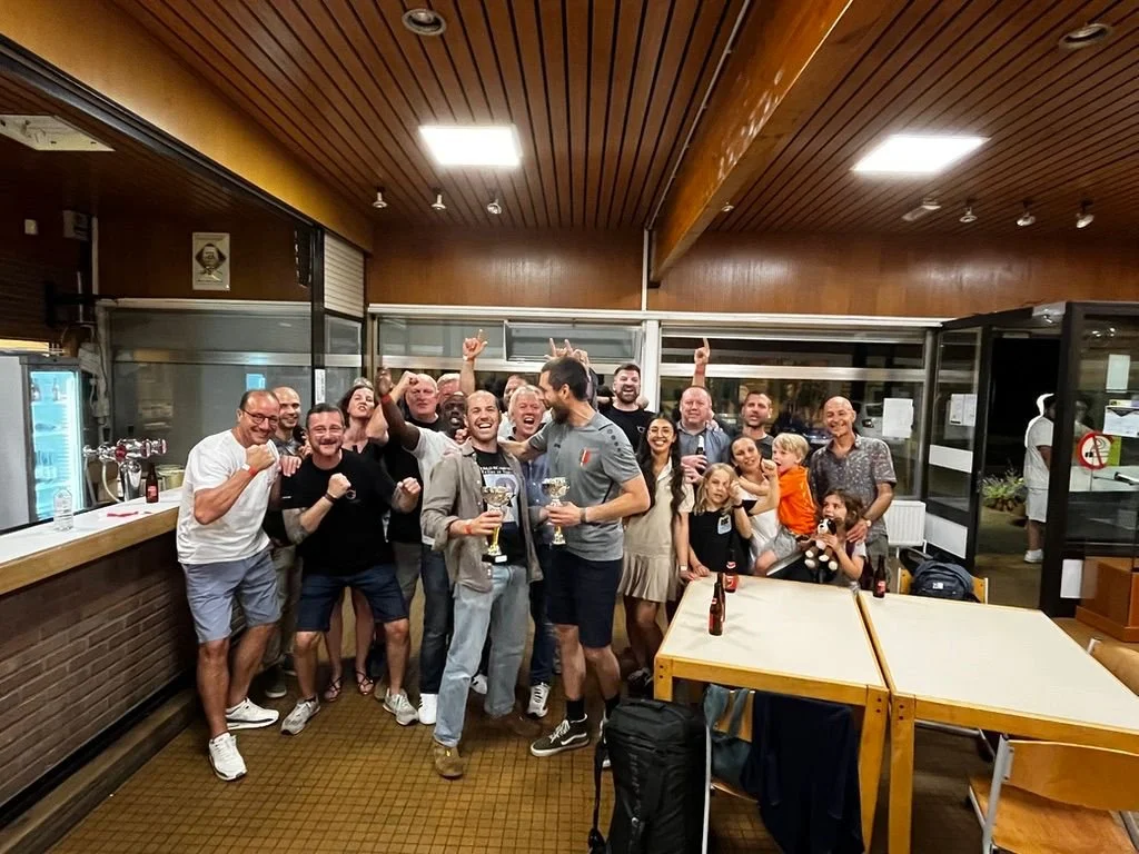 Groupe de personnes souriantes célébrant avec des bières dans un intérieur de restaurant ou de bar en bois, certains avec les bras levés en signe de victoire.