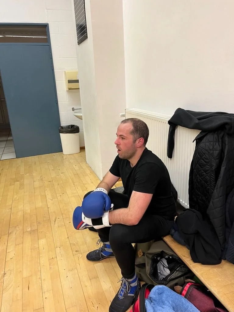 Un homme assis sur un banc dans une salle de sport ou un club de boxe, portant des gants de boxe bleus et habillé en noir, semble fatigué ou concentré.