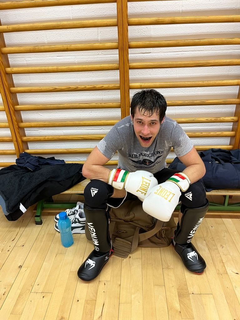 Jeune homme en équipement de boxe avec des gants, assis sur un banc de vestiaire, souriant après un entraînement ou un match.