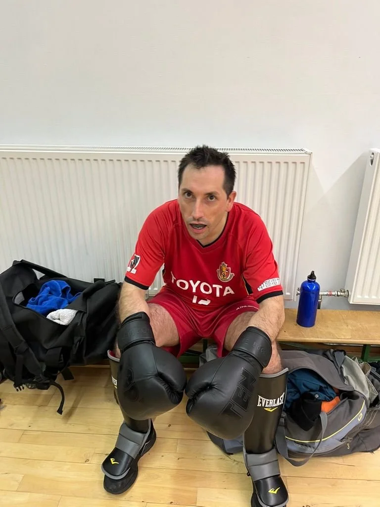 Homme en uniforme de boxe avec gants de boxe noirs, assis dans un vestiaire, semblant se préparer pour un entraînement ou un combat.