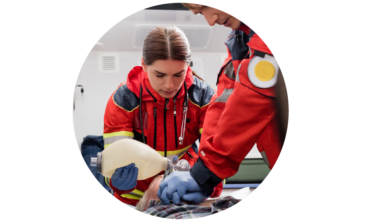Two EMS rescuers wheeling a patient lying on a stretcher into an ambulance. The responder in the foreground is performing CPR chest compressions. The responder in the background is applying an oxygen mask to the patient.