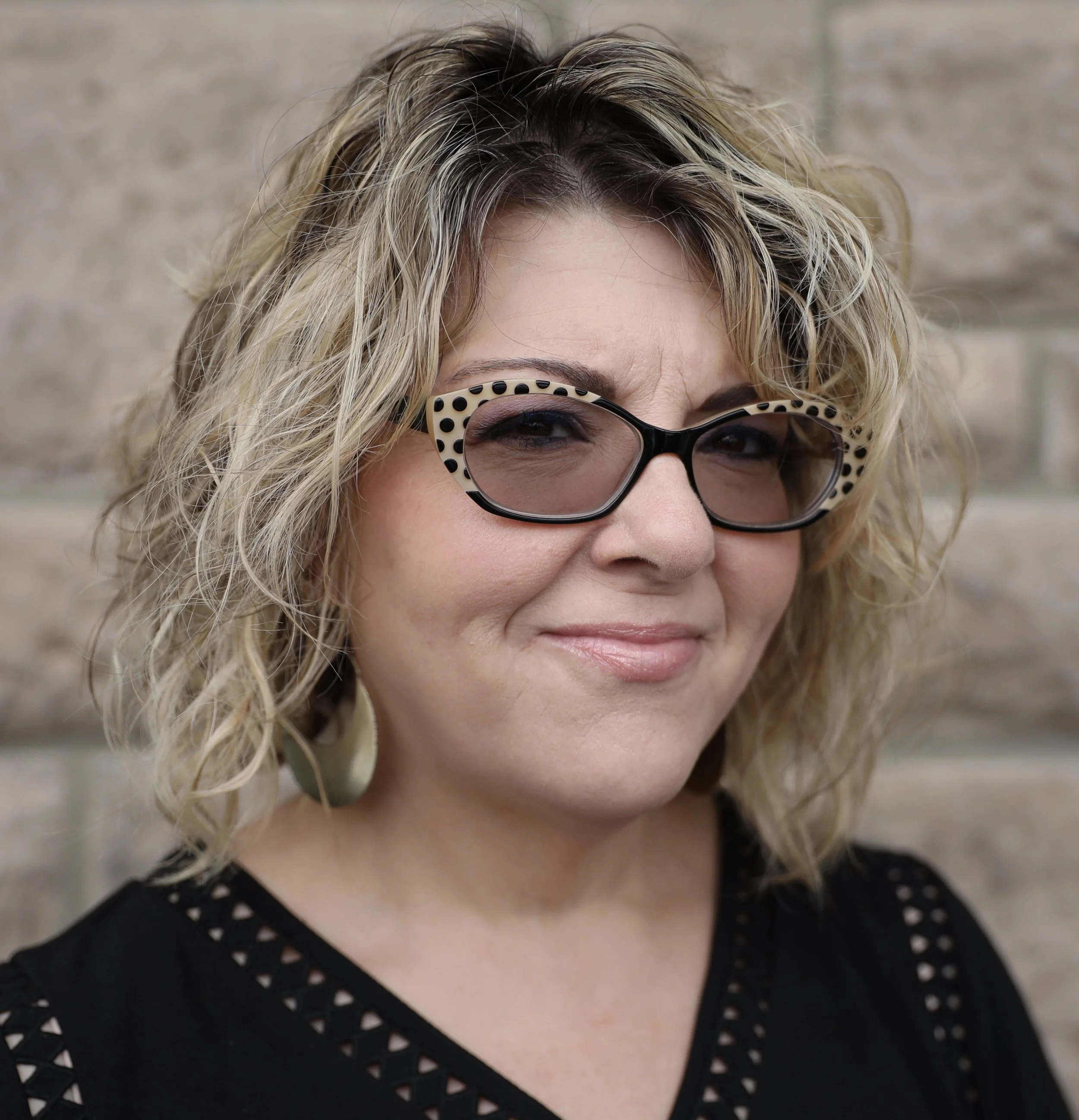 A woman with curly blonde hair wearing black and white polka dot glasses, earrings, and a black top with cut-out details, standing in front of a brick wall.