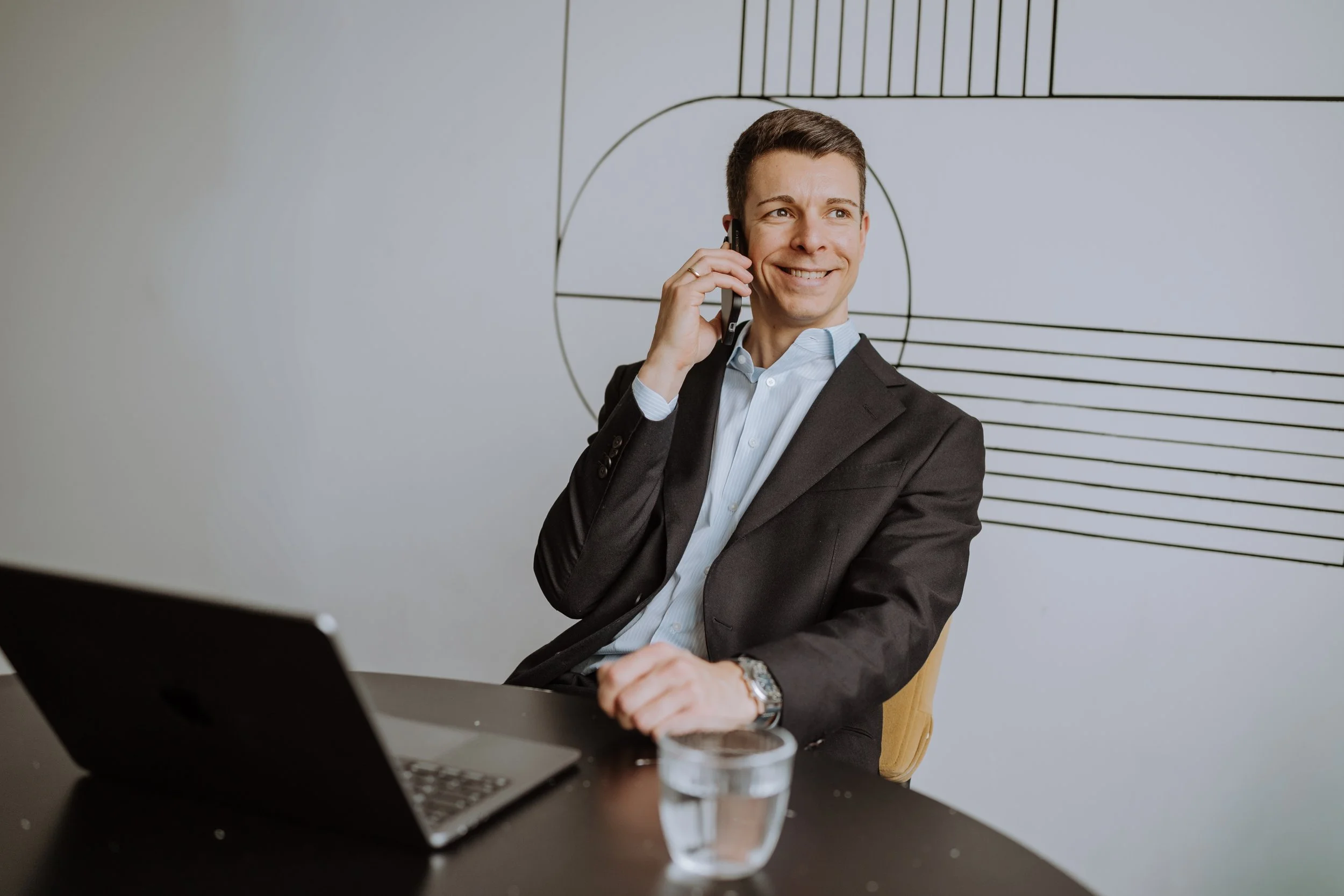 Ein Mann in einem schwarzen Anzug sitzt an einem Tisch, spricht am Telefon, hat einen Laptop vor sich, ein Glas Wasser, und lächelt.