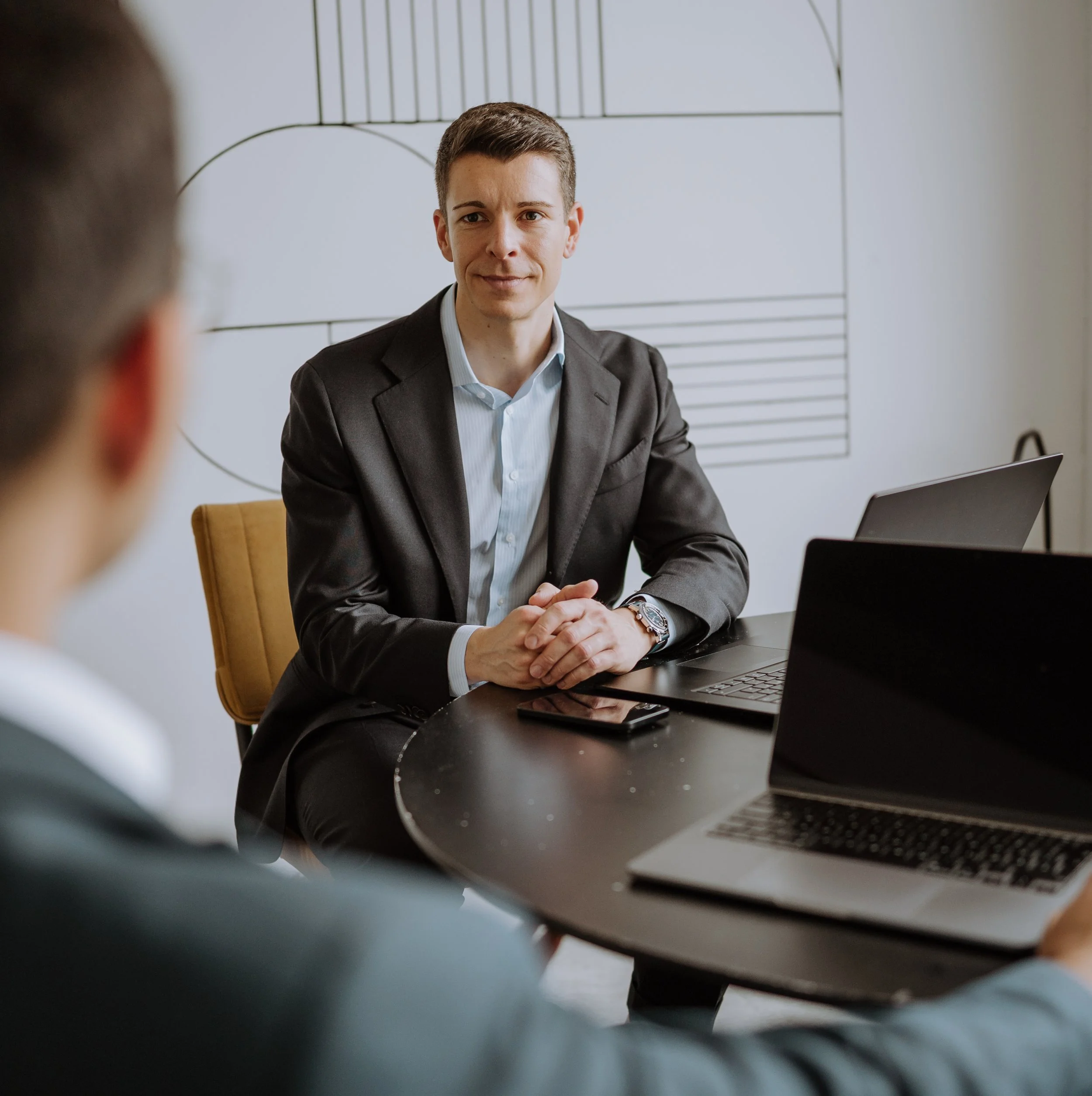 Geschäftsmann im Anzug sitzt an einem Tisch während eines Gesprächs in einem Büro mit Laptops und einem Smartphone.