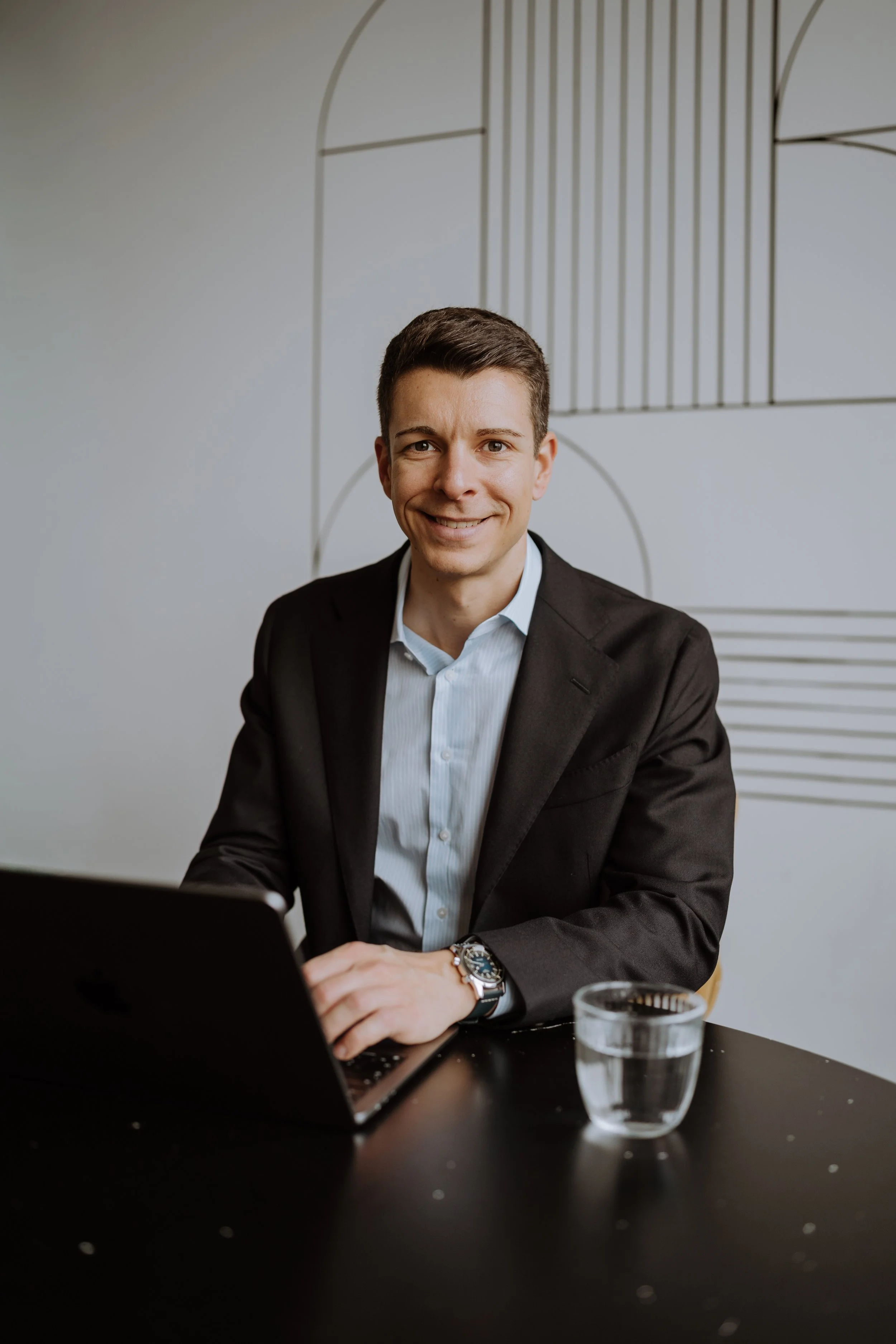 Ein Mann im Anzug sitzt am Tisch mit einem Laptop und einem Glas Wasser, lächelt freundlich, im modernen Büro.