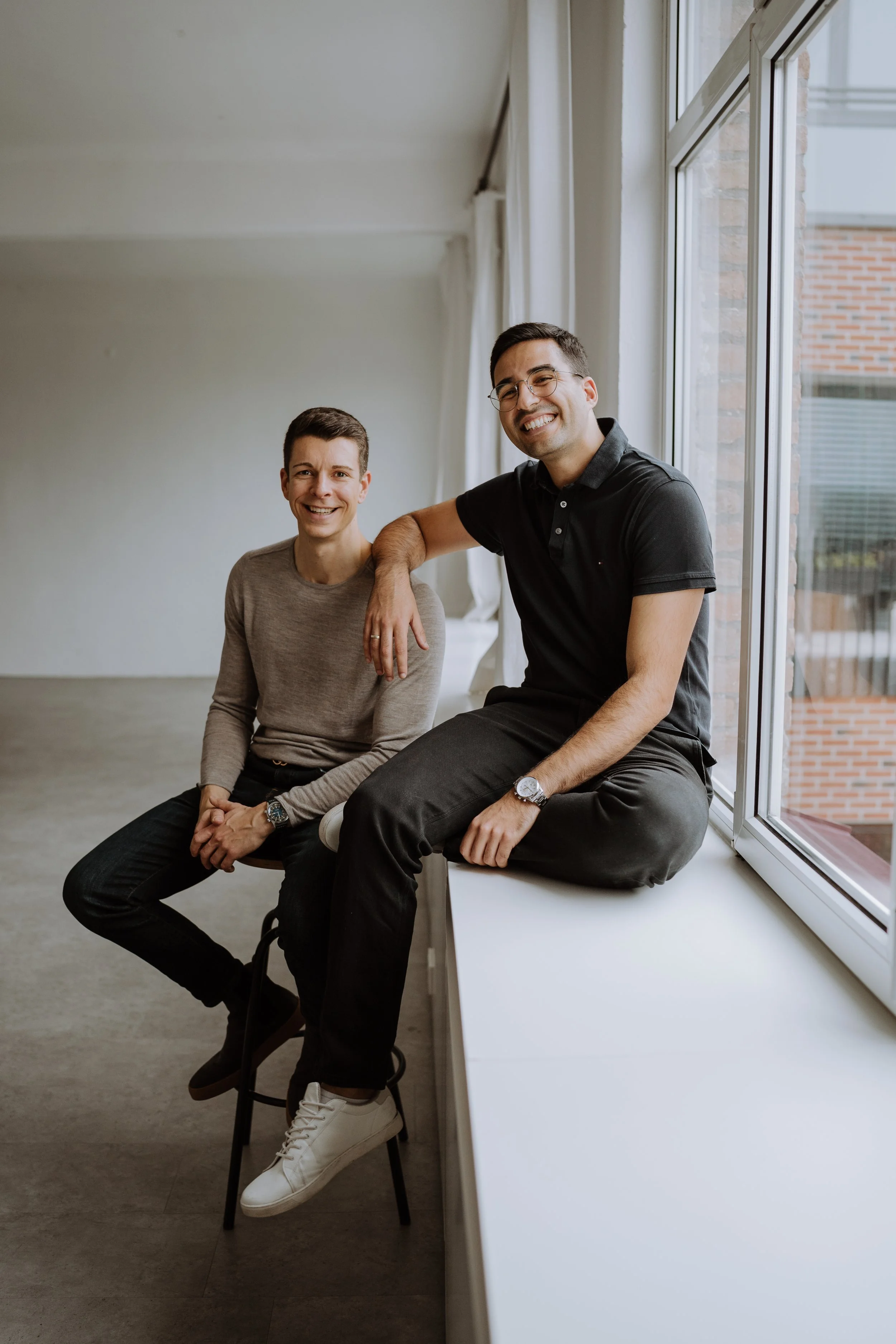 Zwei Männer sitzen und stehen lachend in einem hellen Raum mit großen Fenstern, die Blick auf ein Gebäude mit Backsteinmauer bieten.