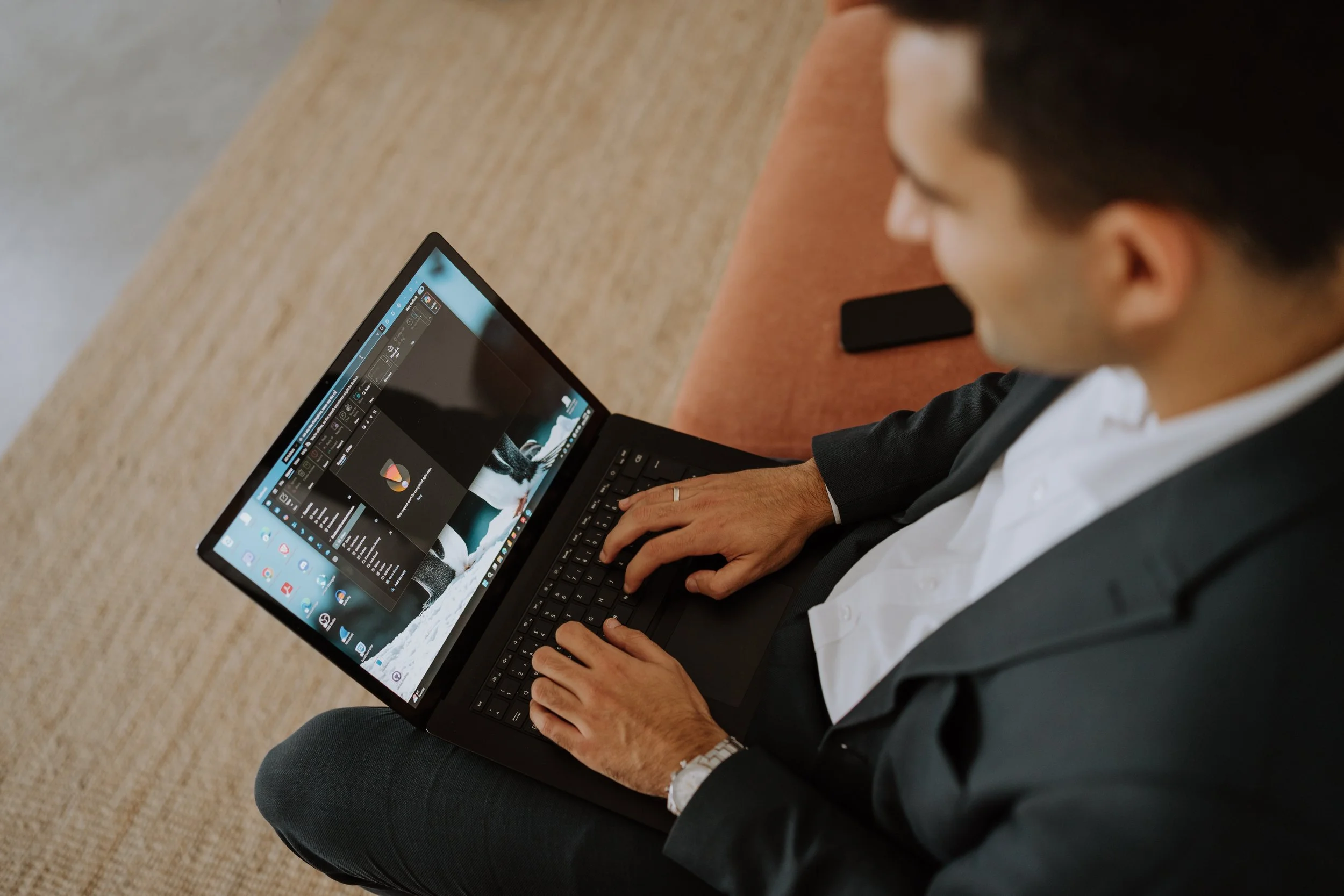 Ein Mann in einem schwarzen Anzug arbeitet an einem Laptop, den er auf seinem Schoß hält, während er auf einem roten Sofa sitzt.