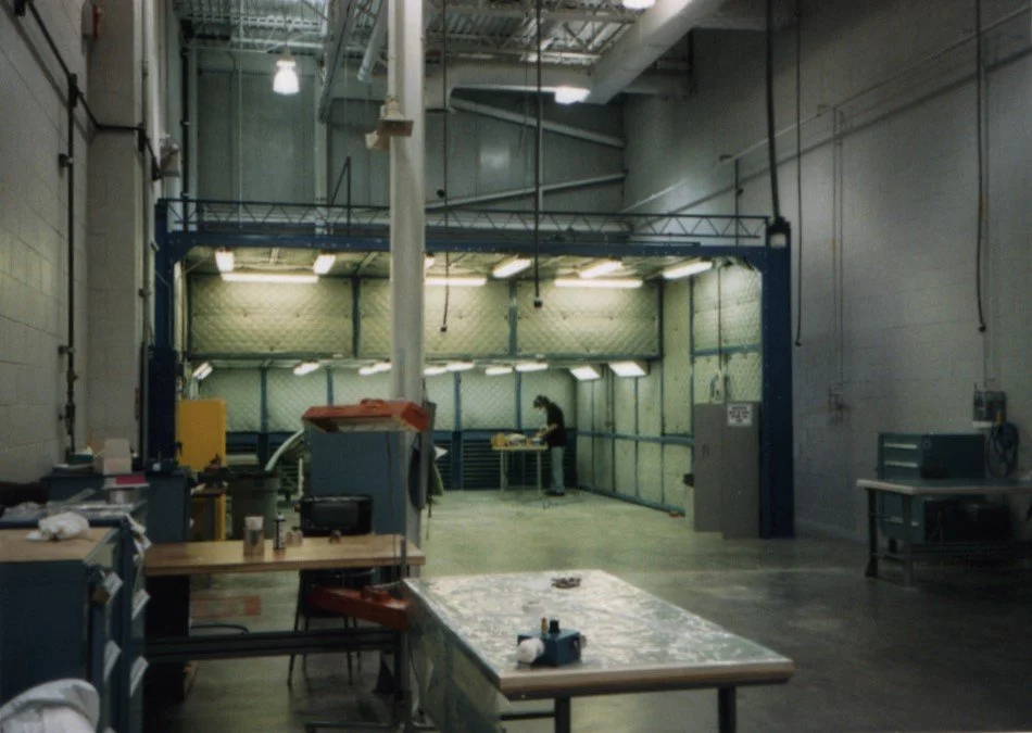 A large industrial workspace or workshop with high ceilings, fluorescent lighting, and various work tables and equipment. A person is working at a station in the background near a fenced area.