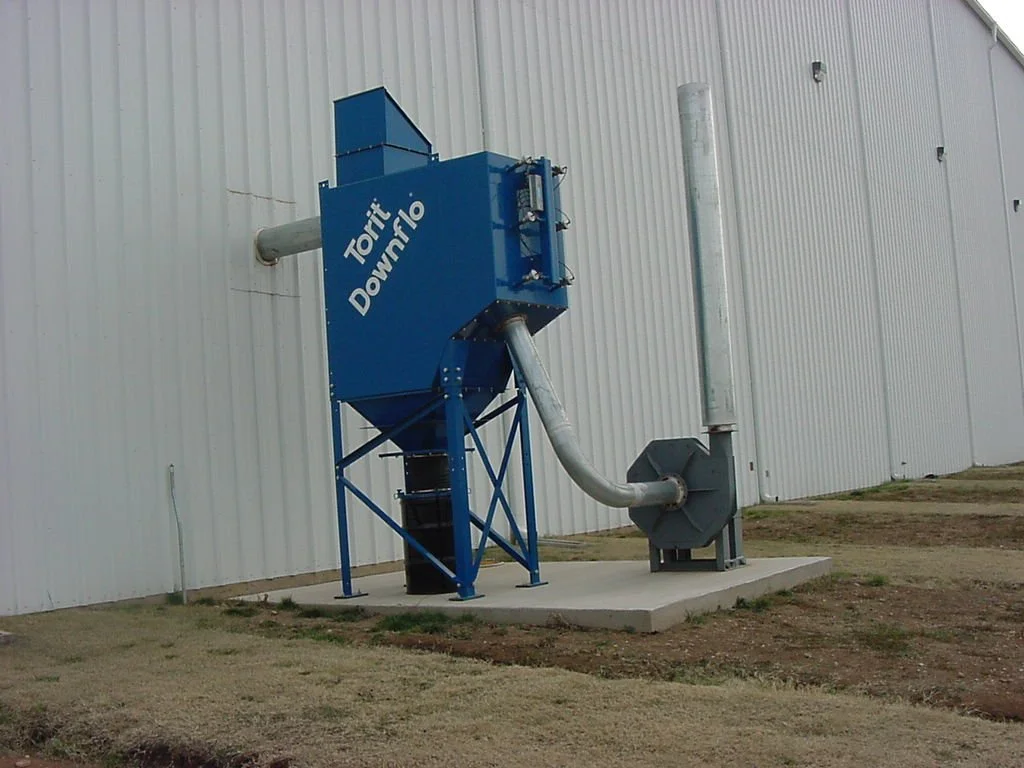 Industrial dust collector system labeled with directions 'To Fit Down' and 'To Regulated' outside a large metal building.