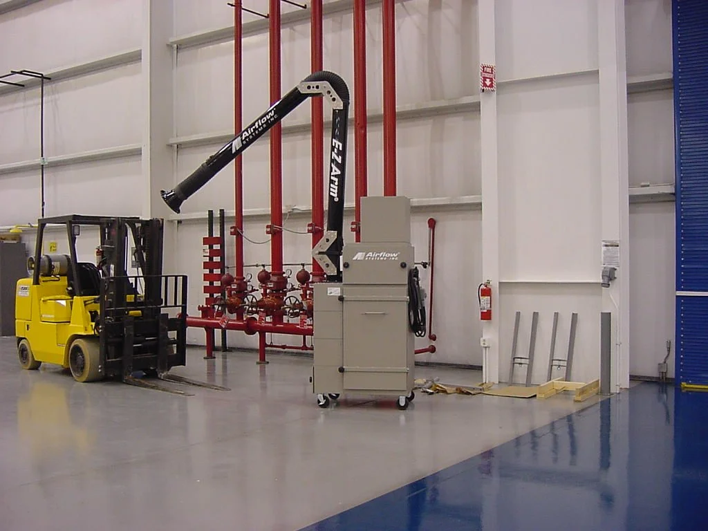 Industrial equipment in a warehouse, including a yellow forklift, a flexible exhaust system, red piping, and a grey storage cabinet with tools nearby.