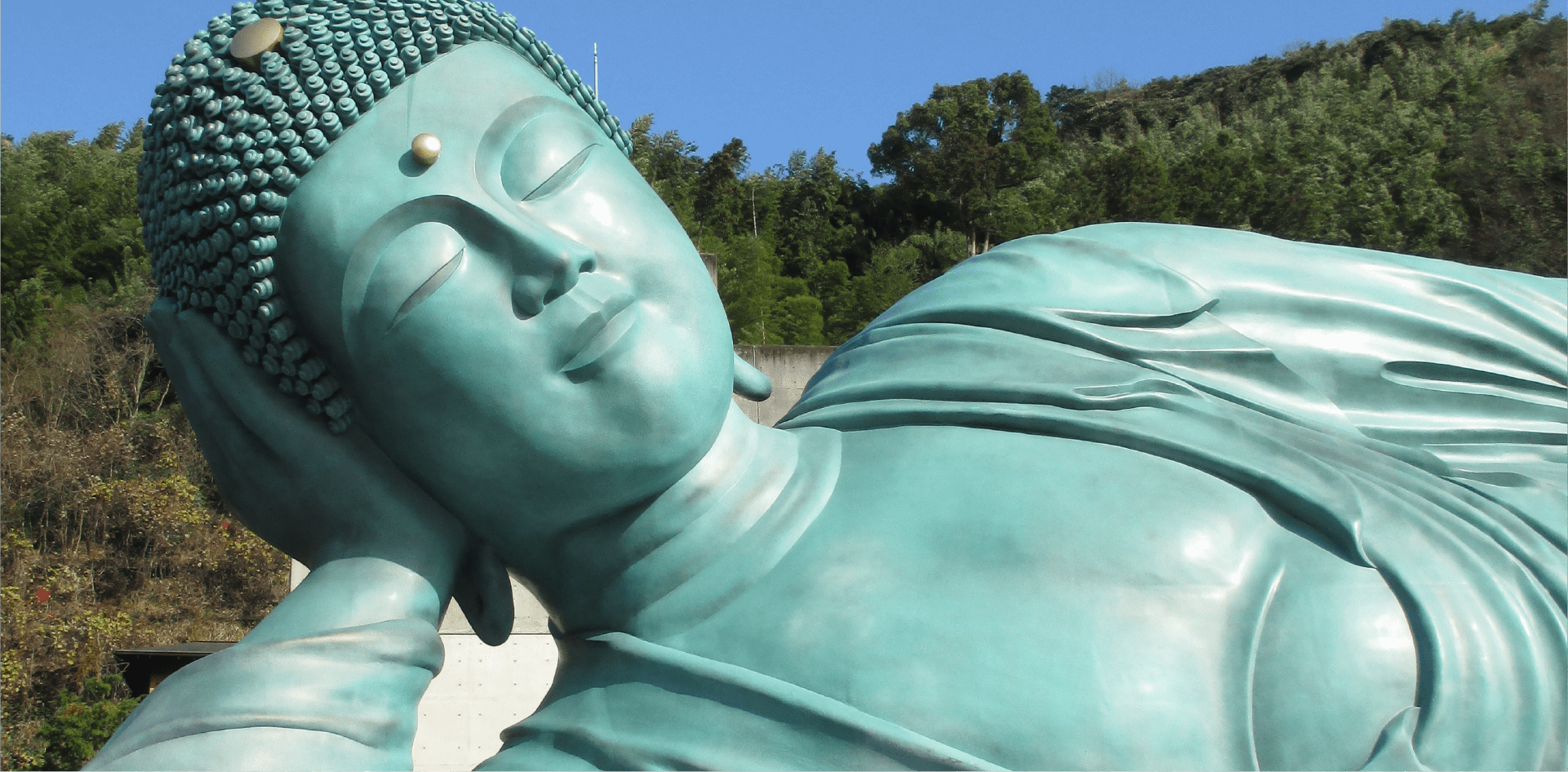 Close-up of the reclining Great Buddha statue made of greenish material, with details of its serene face, hair, and robes, set against a background of blue sky and green hills.
