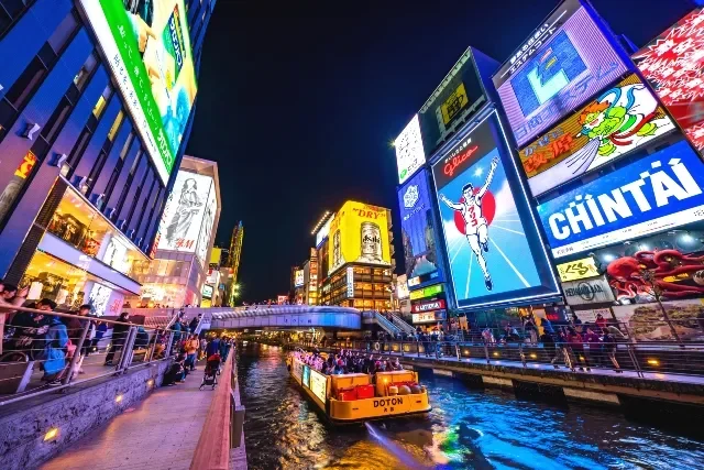 Brightly lit buildings and billboards along a canal at night, with a boat named Doton sailing through, in a lively urban area.