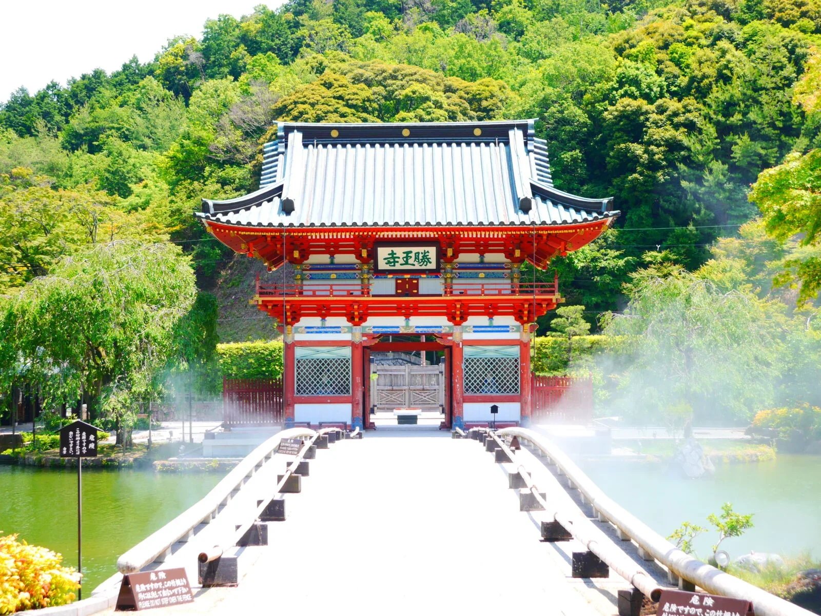 Traditional Japanese temple with red and white architecture, situated over a pond with a bridge leading to the entrance, surrounded by lush green trees.