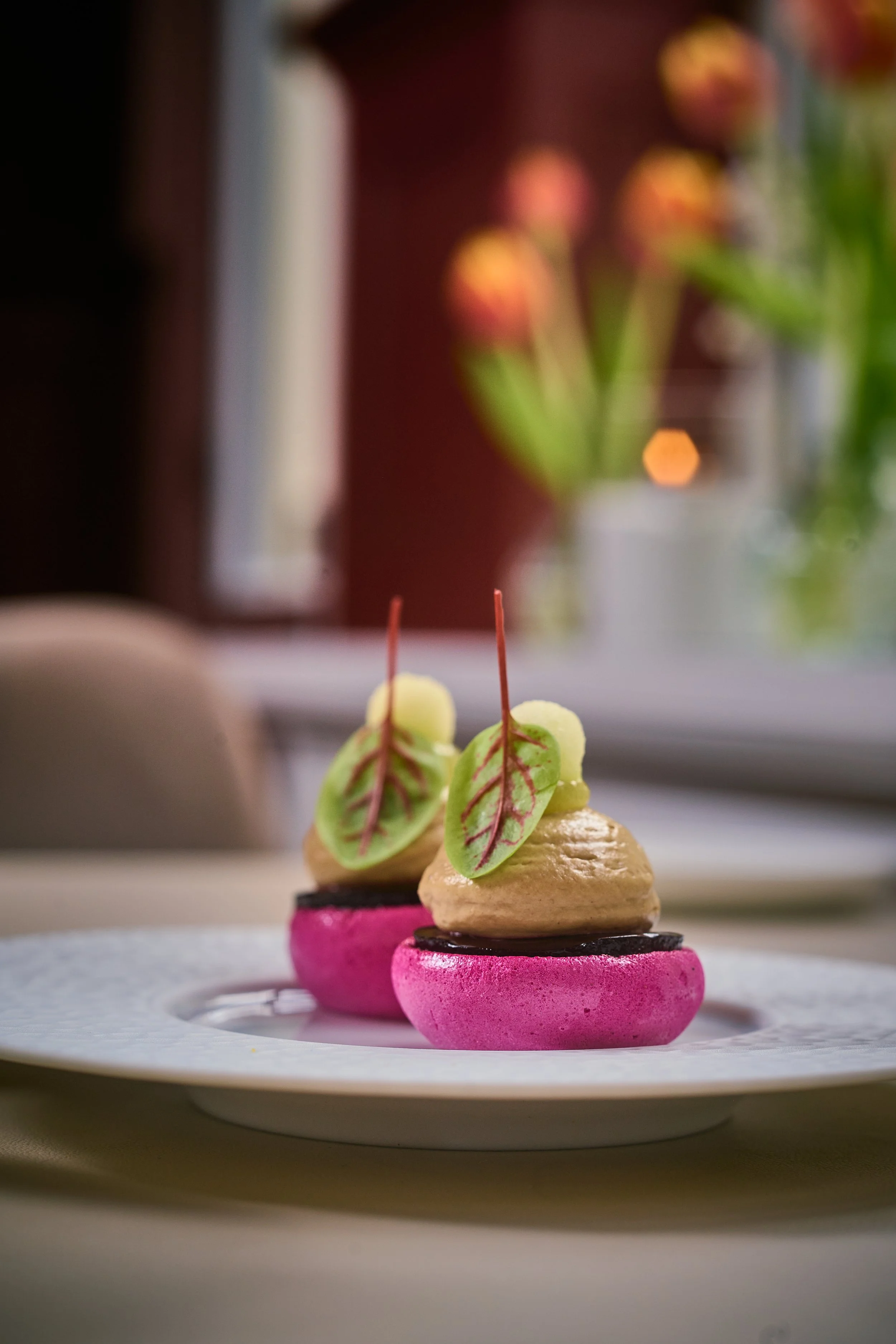 Een zomerse amuse van Restaurant Blumé, ook wel mondvermaakje, die zintuigen prikkelt. Roze macarons, groene decoratieve bladeren en crème, geserveerd op een wit bord.