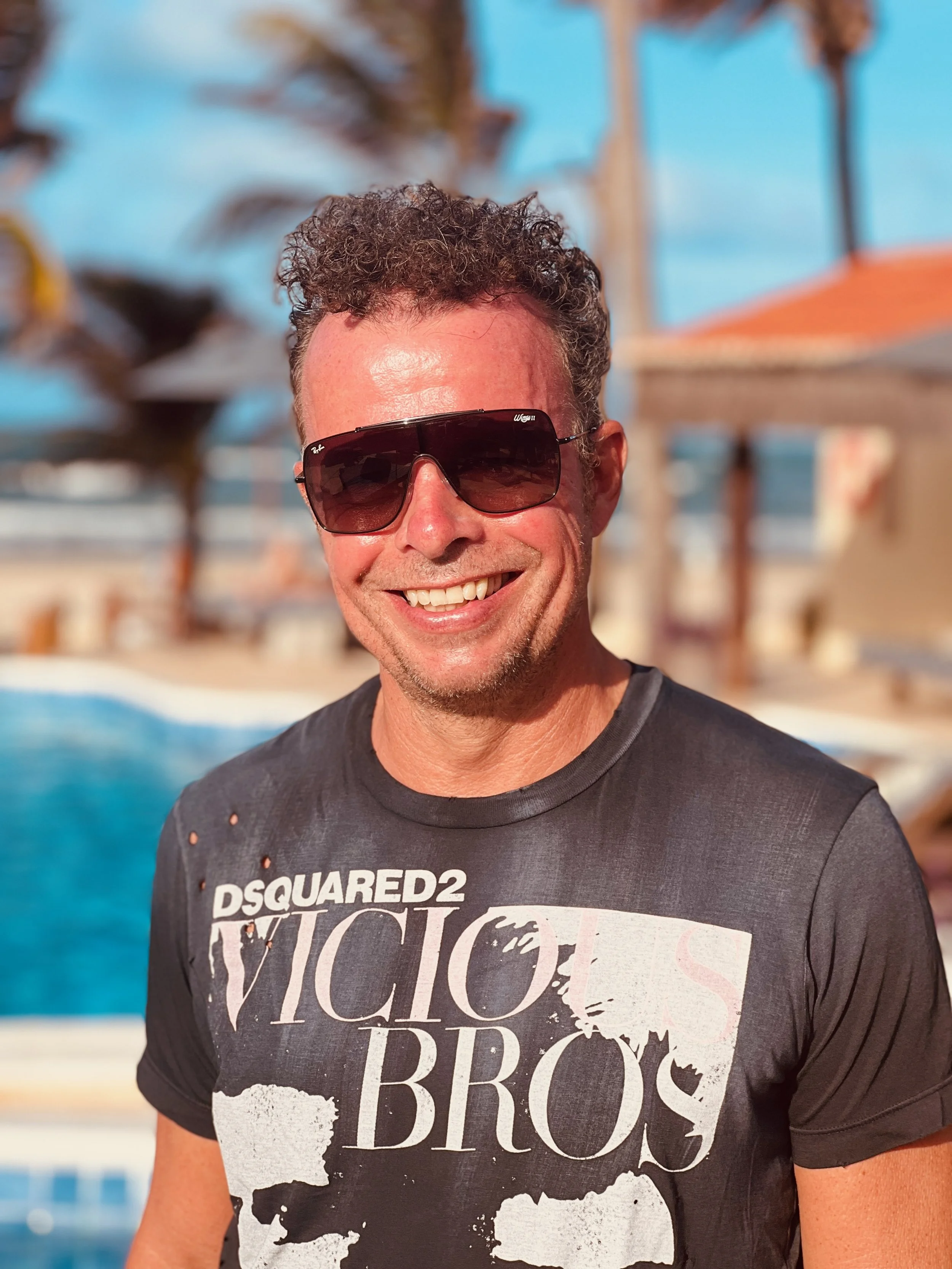 Lächelnder Mann mit dunklen Sonnenbrillen und einem grauen T-Shirt vor einem Pool und Palmen im Hintergrund bei Sonnenlicht.