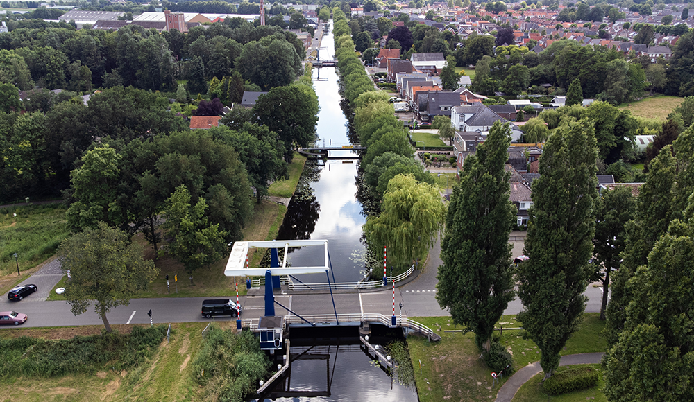 Luchtfoto van een kanaal met een sluis, omgeven door groene bomen en een woonwijk met huizen en wegen.