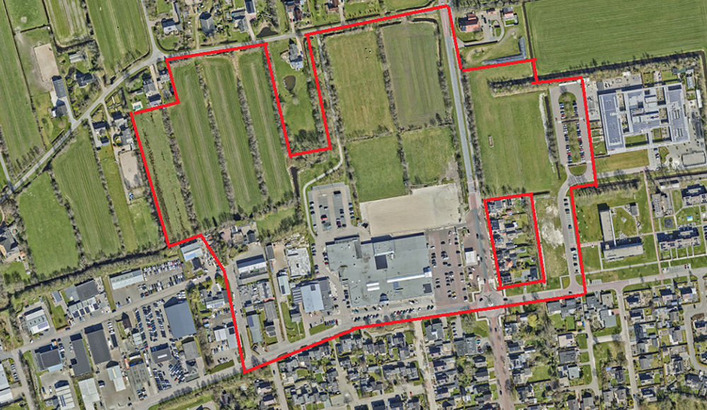 Luchtfoto van een gebied met velden, wegen, gebouwen en parkeerplaatsen, met een rode omtreklijn rond een deel van het terrein.