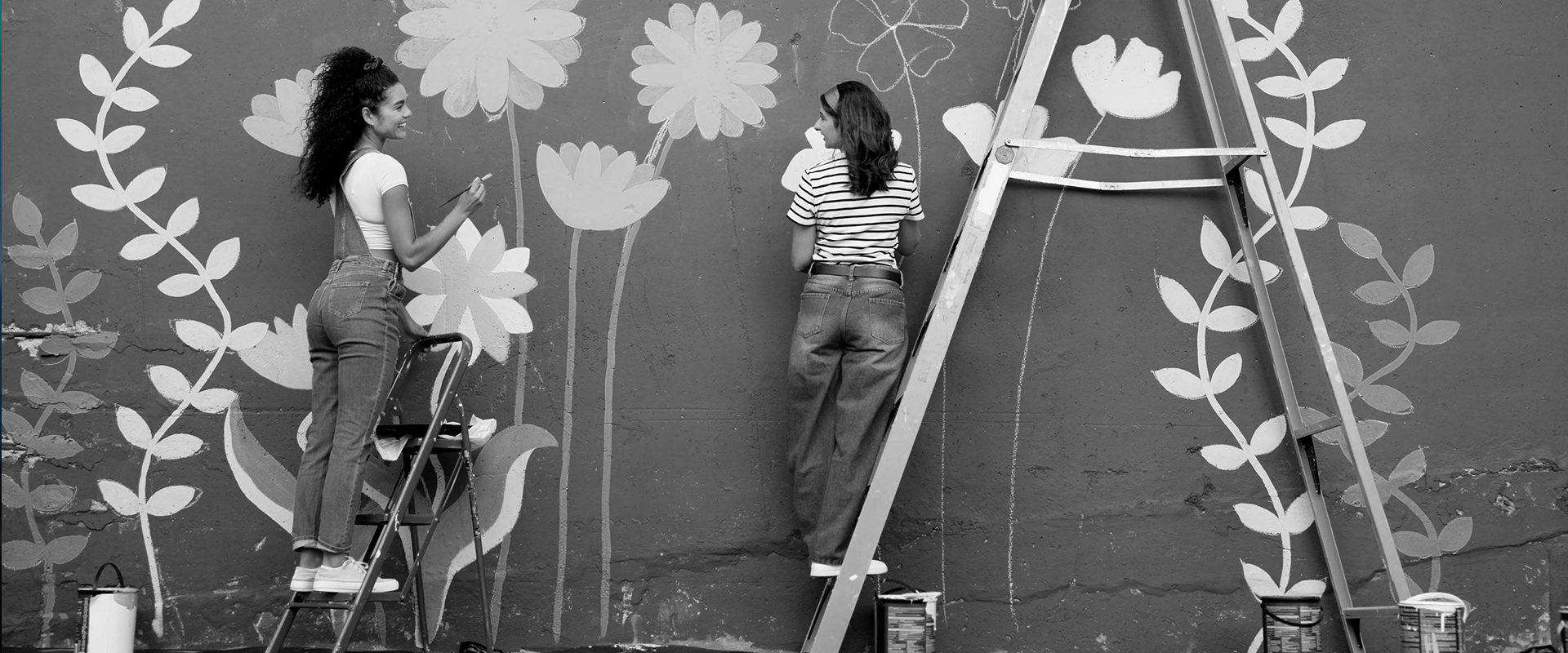 Twee vrouwen schilderen een muurschildering met bloemen op een muur, ondersteund door een ladder en verfblikken.