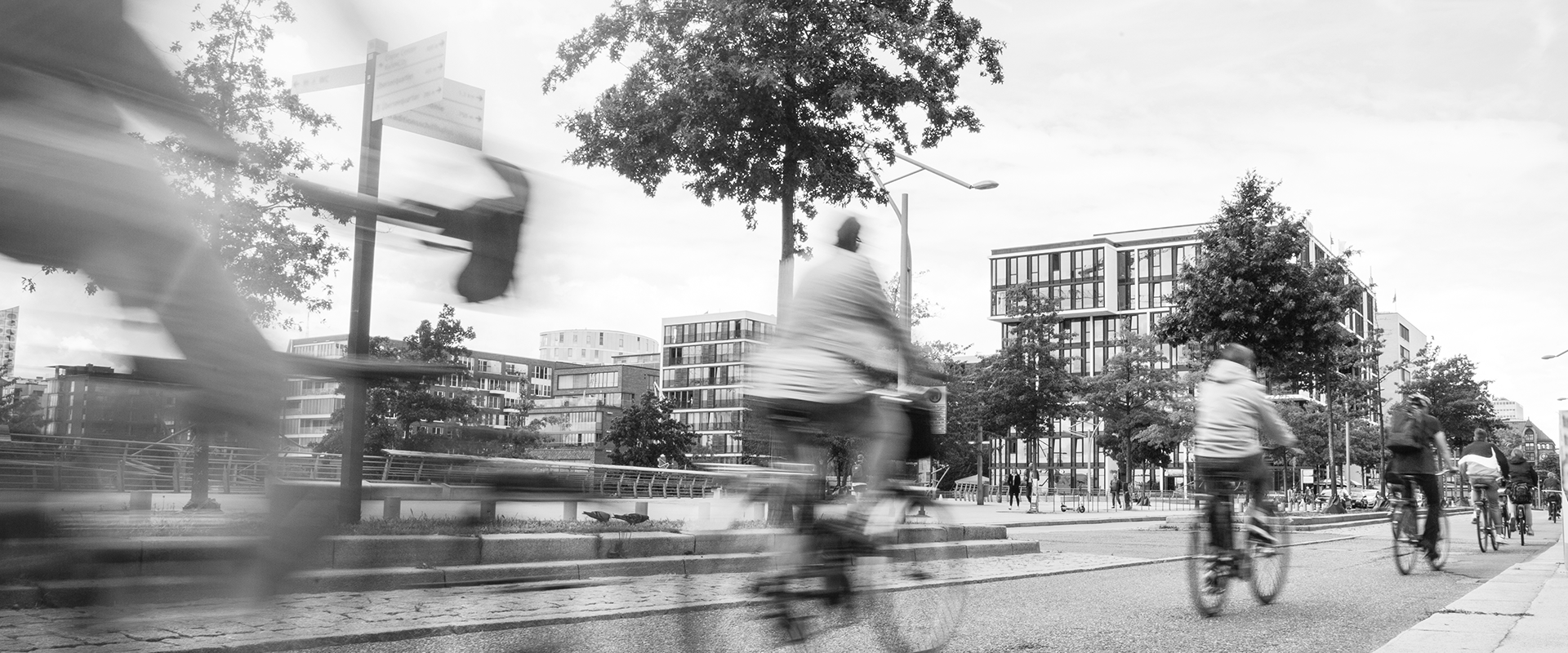 Fietsers rijden door een stedelijk gebied met moderne gebouwen en bomen, terwijl een paar mensen te voet lopen op de achtergrond.