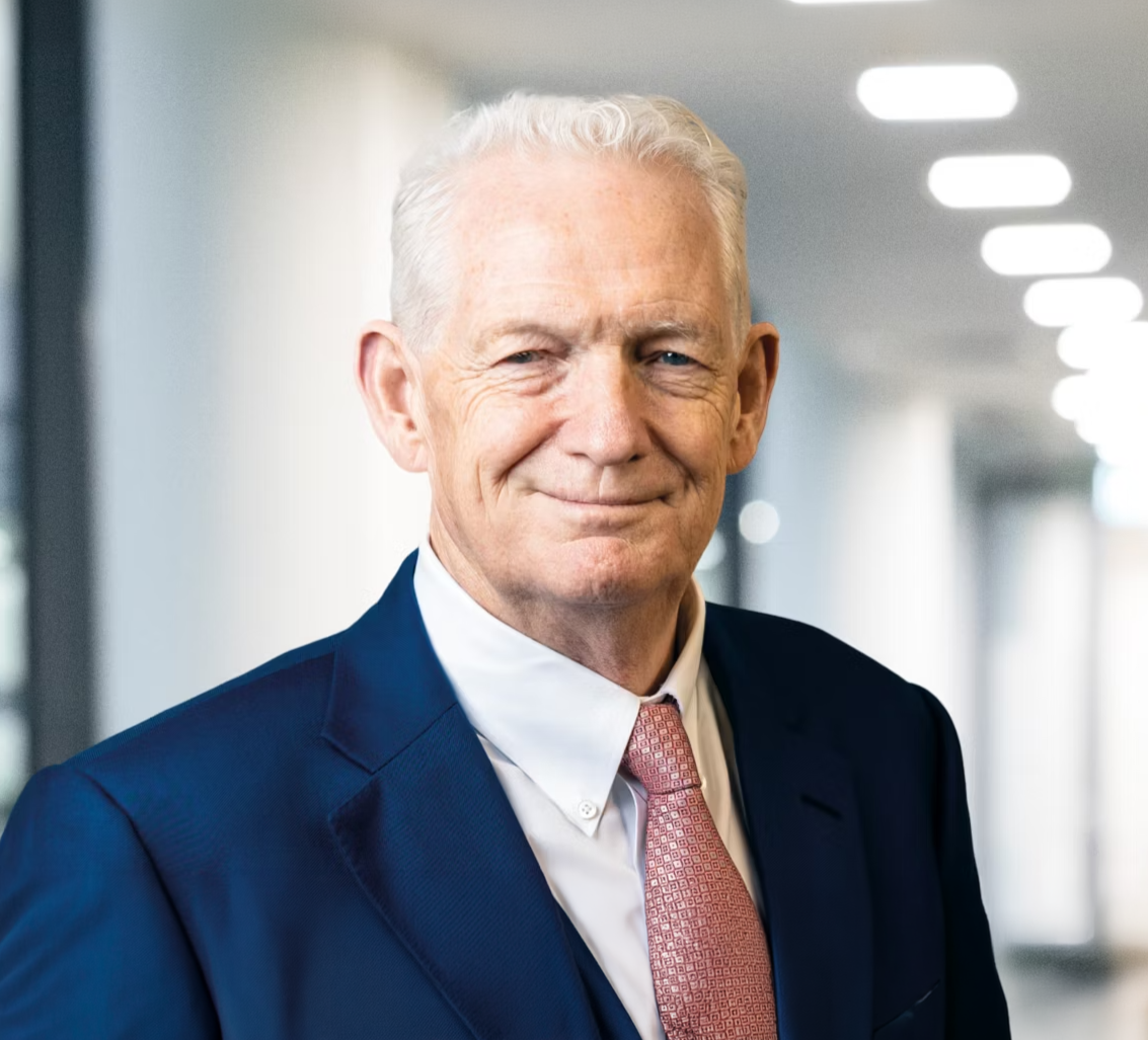 A portrait of an older man with white hair, dressed in a navy suit, white shirt, and pink tie, standing in a modern office hallway.