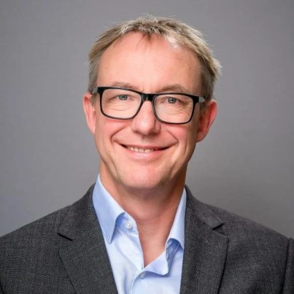 A middle-aged man with light brown hair, wearing glasses, a light blue shirt, and a dark gray blazer, smiling against a gray background.