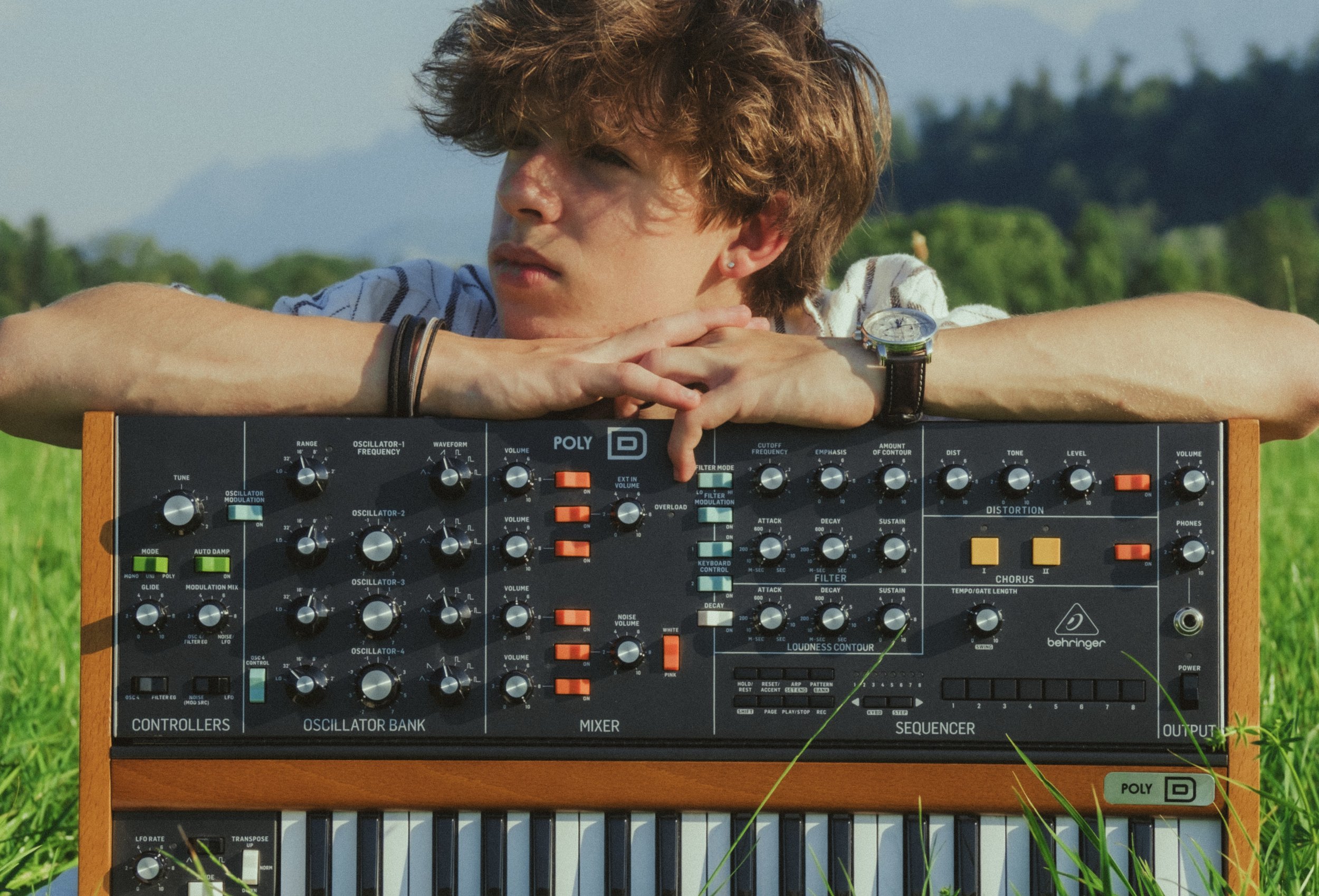 Young man with brown shaggy hair resting his chin on a vintage synthesizer outdoors in a grassy field, with mountains and trees in the background.