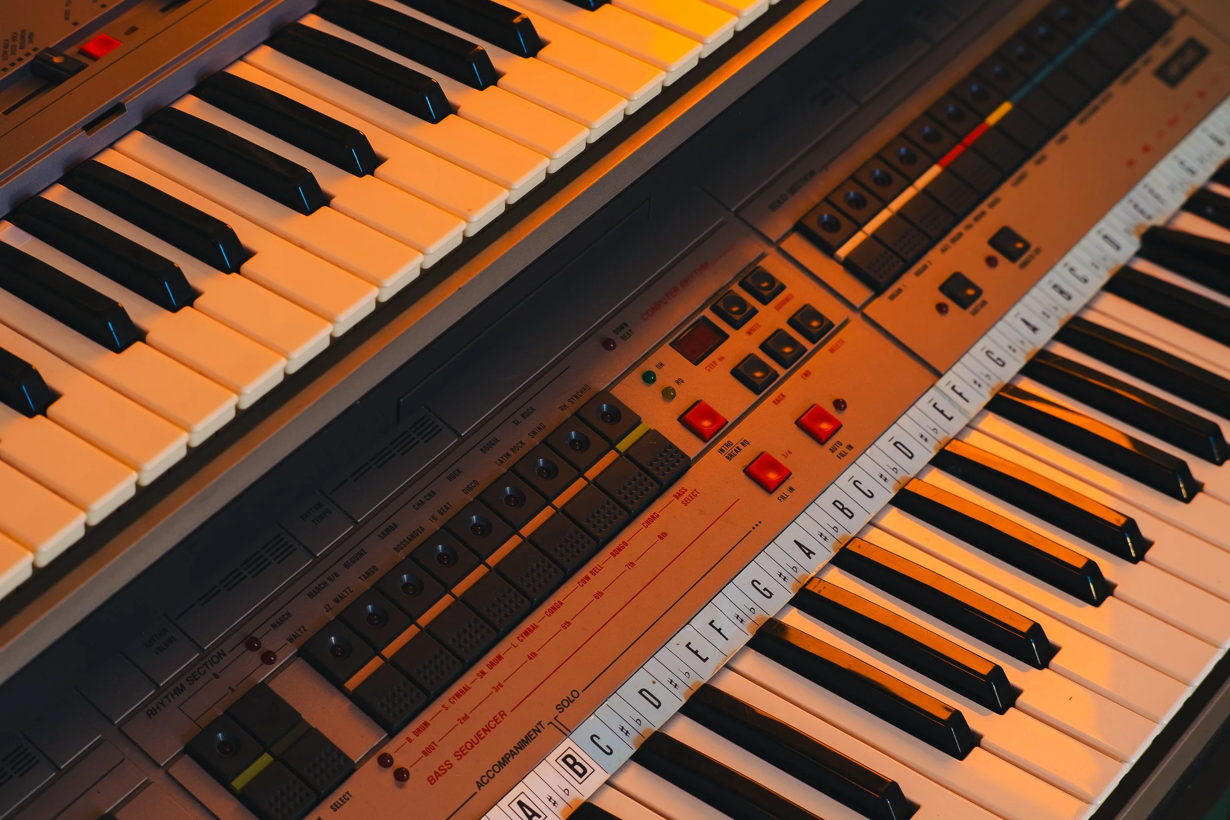 Close-up of a vintage electronic keyboard synthesizer with black and white keys, various control buttons, and colorful sliders.