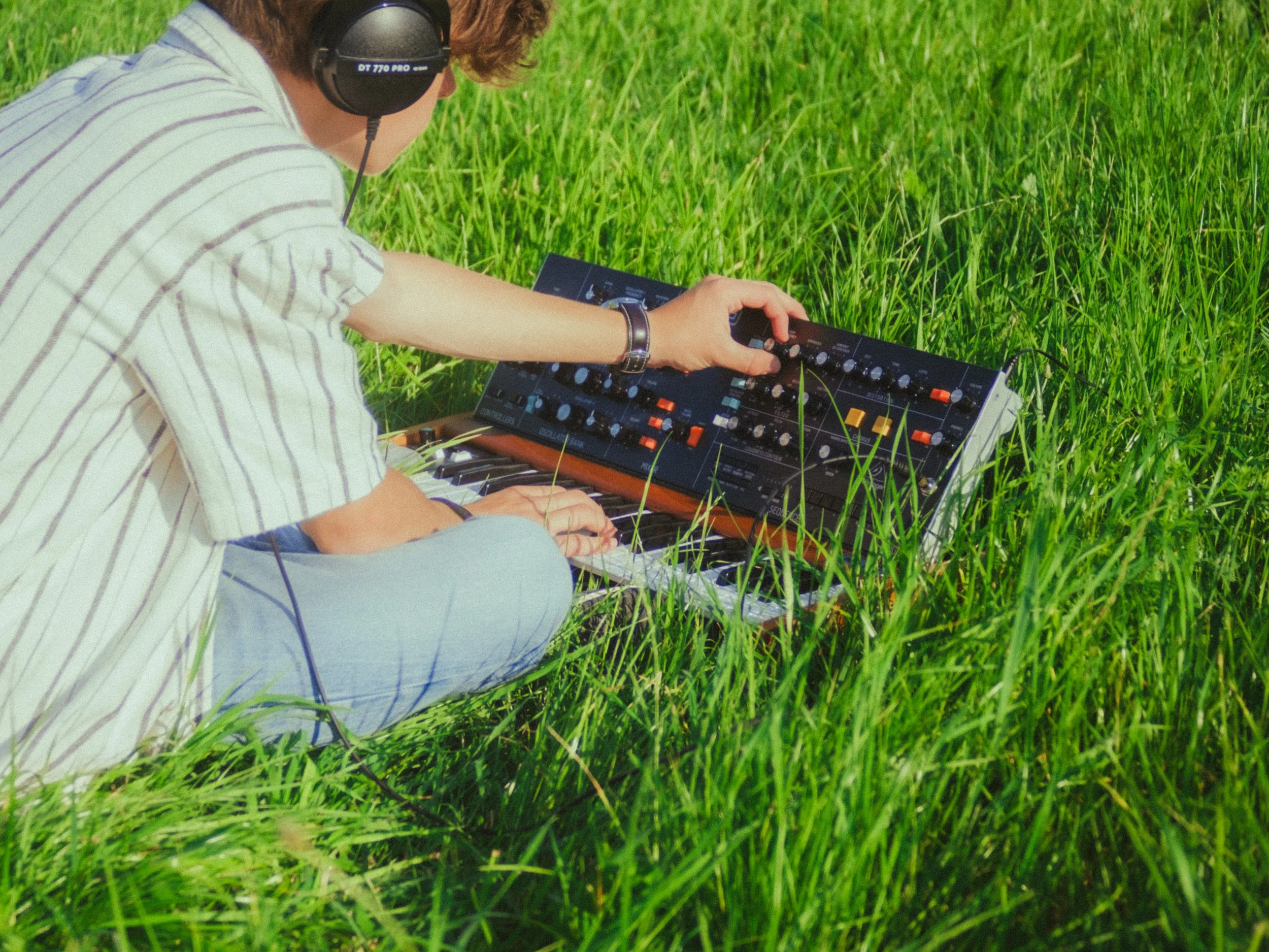 A person sitting in a grassy field wearing headphones, adjusting a synthesizer.
