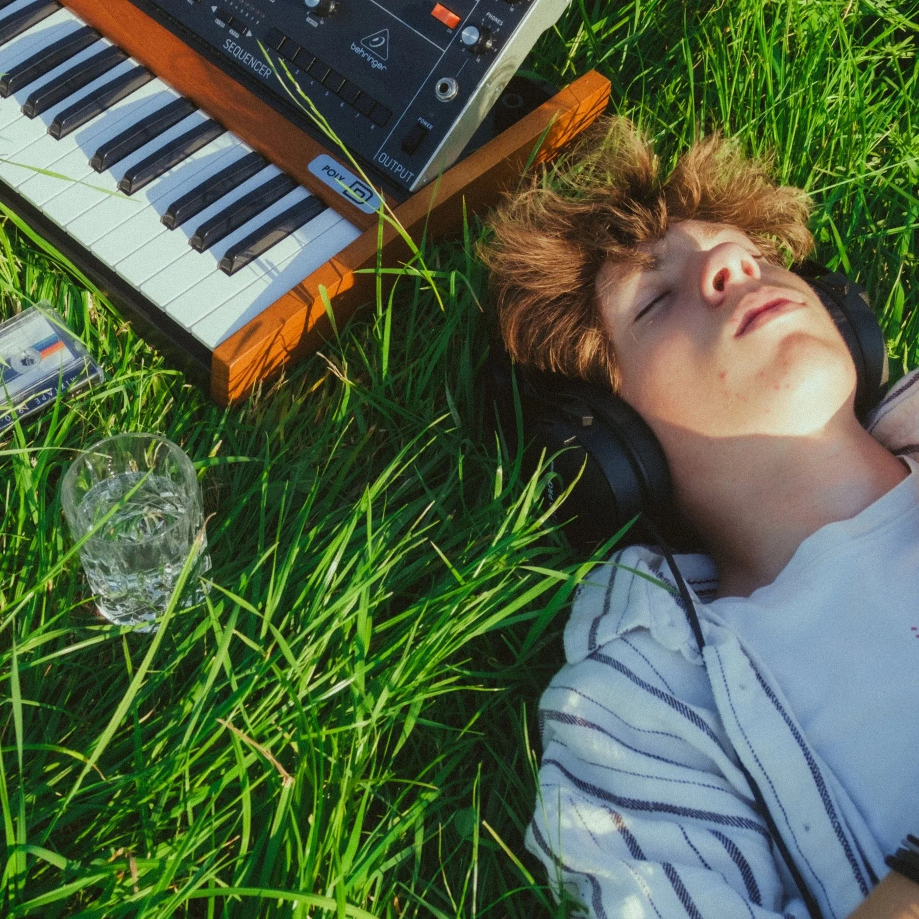 A young person with headphones lying on grass with eyes closed, near a vintage keyboard and audio equipment, on a sunny day.