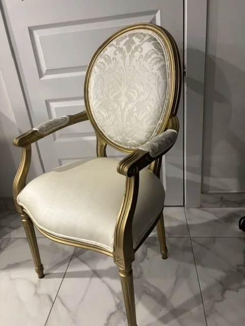 A vintage-style armchair with a gold frame, light patterned upholstery on the backrest, plain cream fabric on the seat, and padded armrests, placed on a marble floor in front of a white paneled door.